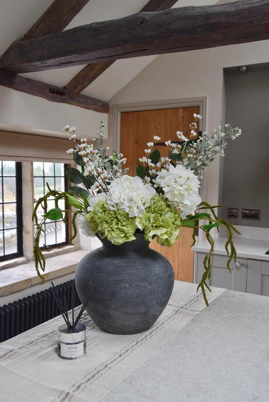 Grey vase placed on a kitchen countertop with a mixture of green and white hydrangeas with lots of mixed foliage. Displayed in a country style kitchen for home decor.