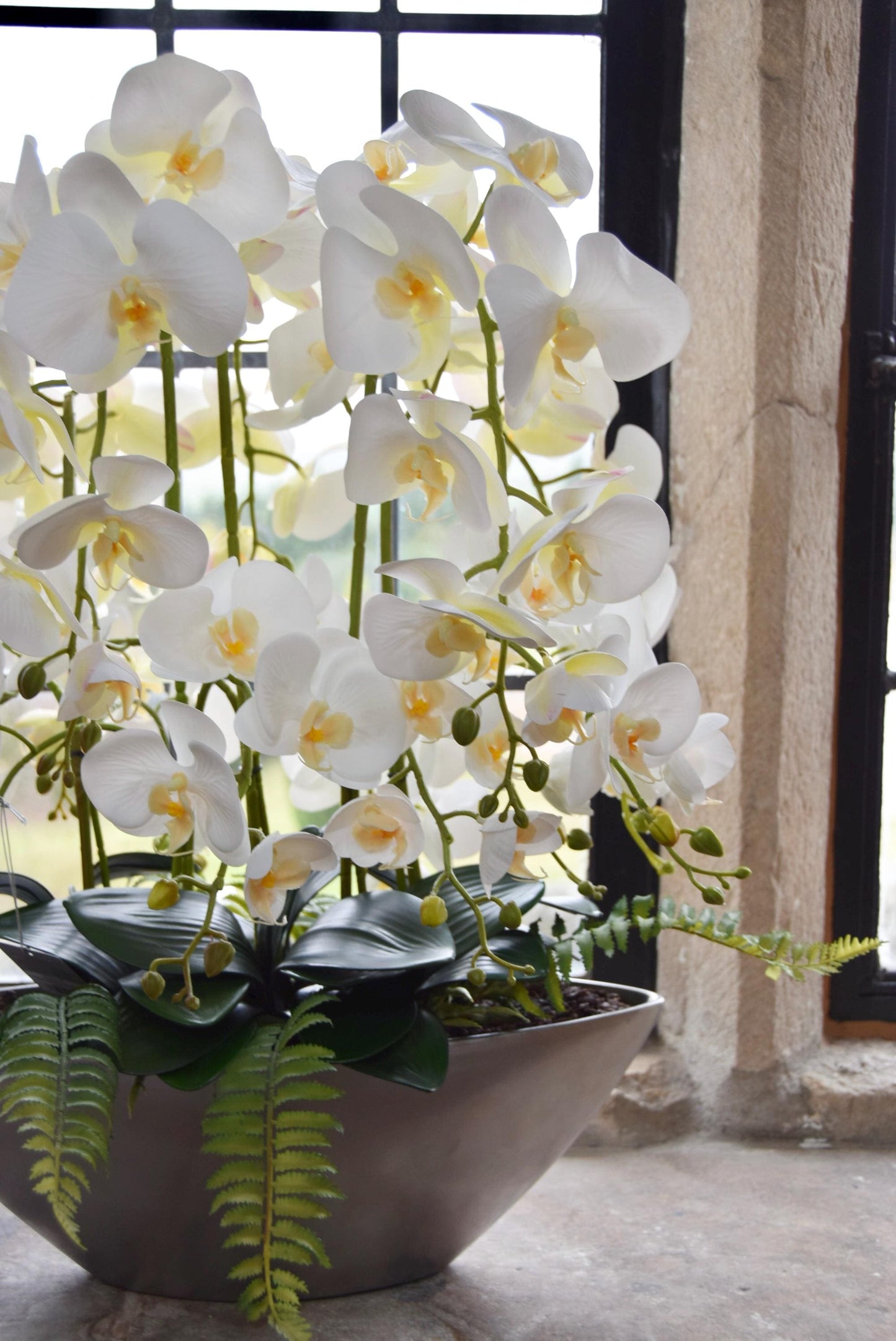 Silver ark bowl placed on a stone windowsill with an arrangement of white orchids for home decor. Arranged in a country style kitchen for home decor.