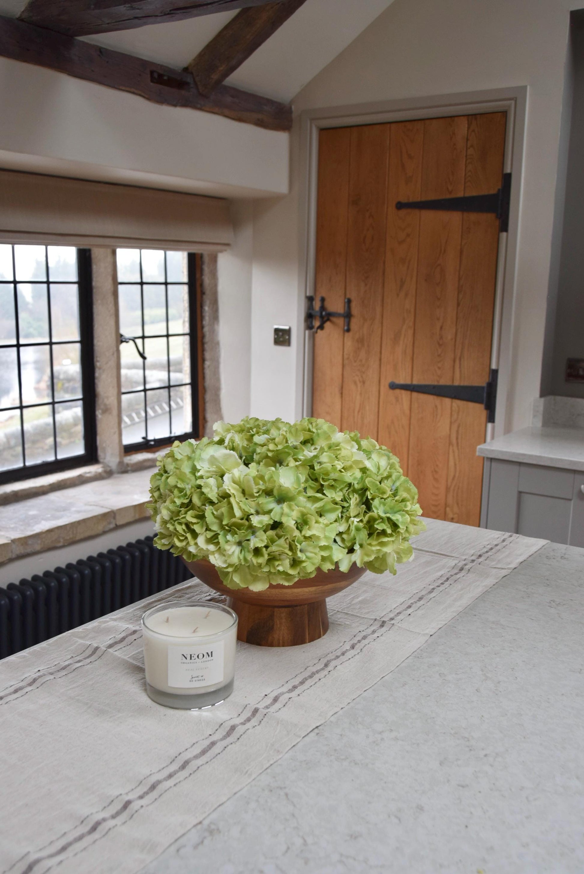 Wooden footed chalice bowl placed on a kitchen counter top with an arrangement of green hydrangeas. Displayed in a country style kitchen for home decor.