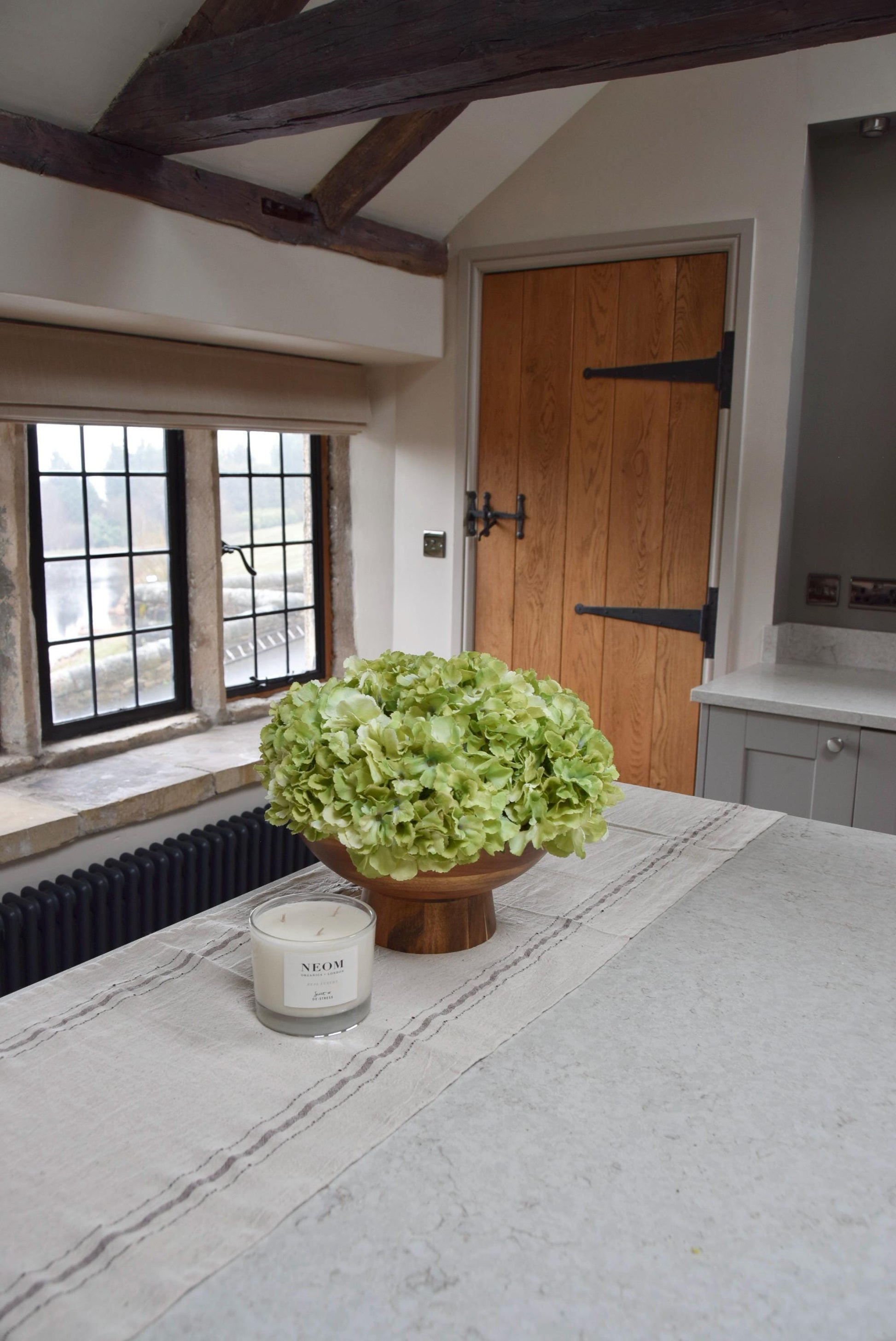 Wooden footed chalice bowl placed on a kitchen counter top with an arrangement of green hydrangeas. Displayed in a country style kitchen for home decor.