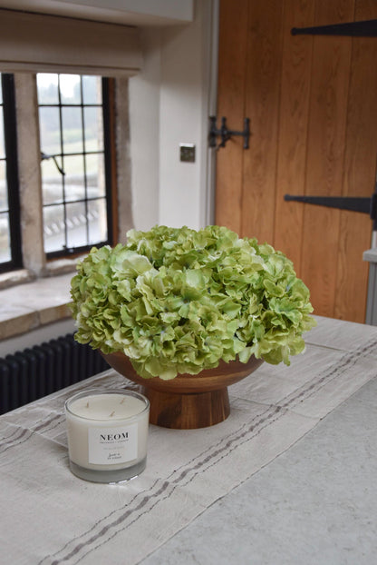 Wooden footed chalice bowl placed on a kitchen counter top with an arrangement of green hydrangeas. Displayed in a country style kitchen for home decor.