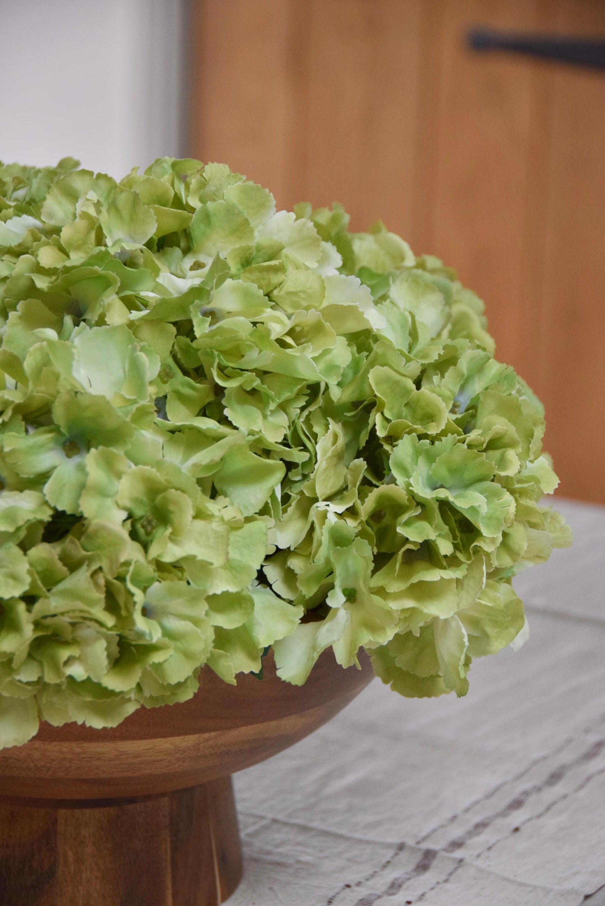 Wooden footed chalice bowl placed on a kitchen counter top with an arrangement of green hydrangeas. Displayed in a country style kitchen for home decor.