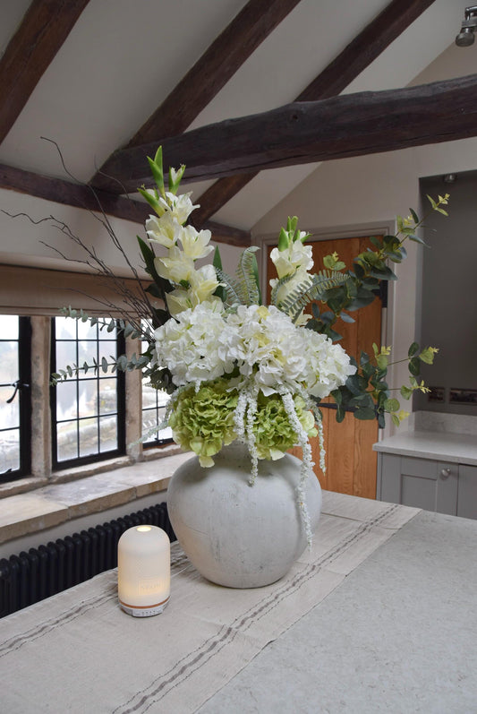 White stone vase placed on a kitchen countertop with a mixture of green hydrangeas, white hydrangeas, gladioli and green foliage. Displayed in a country style kitchen for home decor.