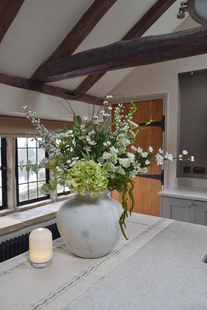 White stone vase placed on a kitchen countertop with a mixture of green hydrangeas, white cherry blossoms and white bud sprays with foliage. Displayed in a country style kitchen for home decor.