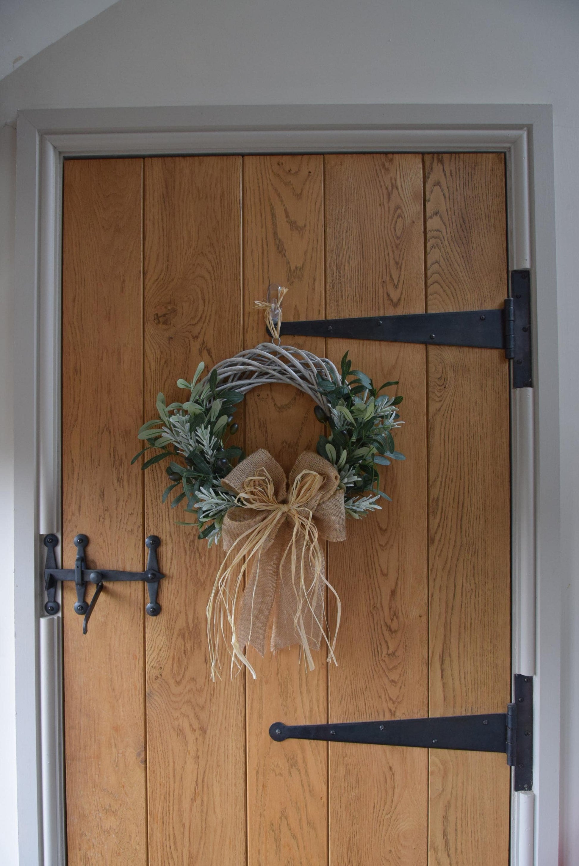 Olive branch wreath created on a willow wreath hanging on a country style door. Mixed with olive branches and tarragon herbs, finished with raffia and natural ribbon.