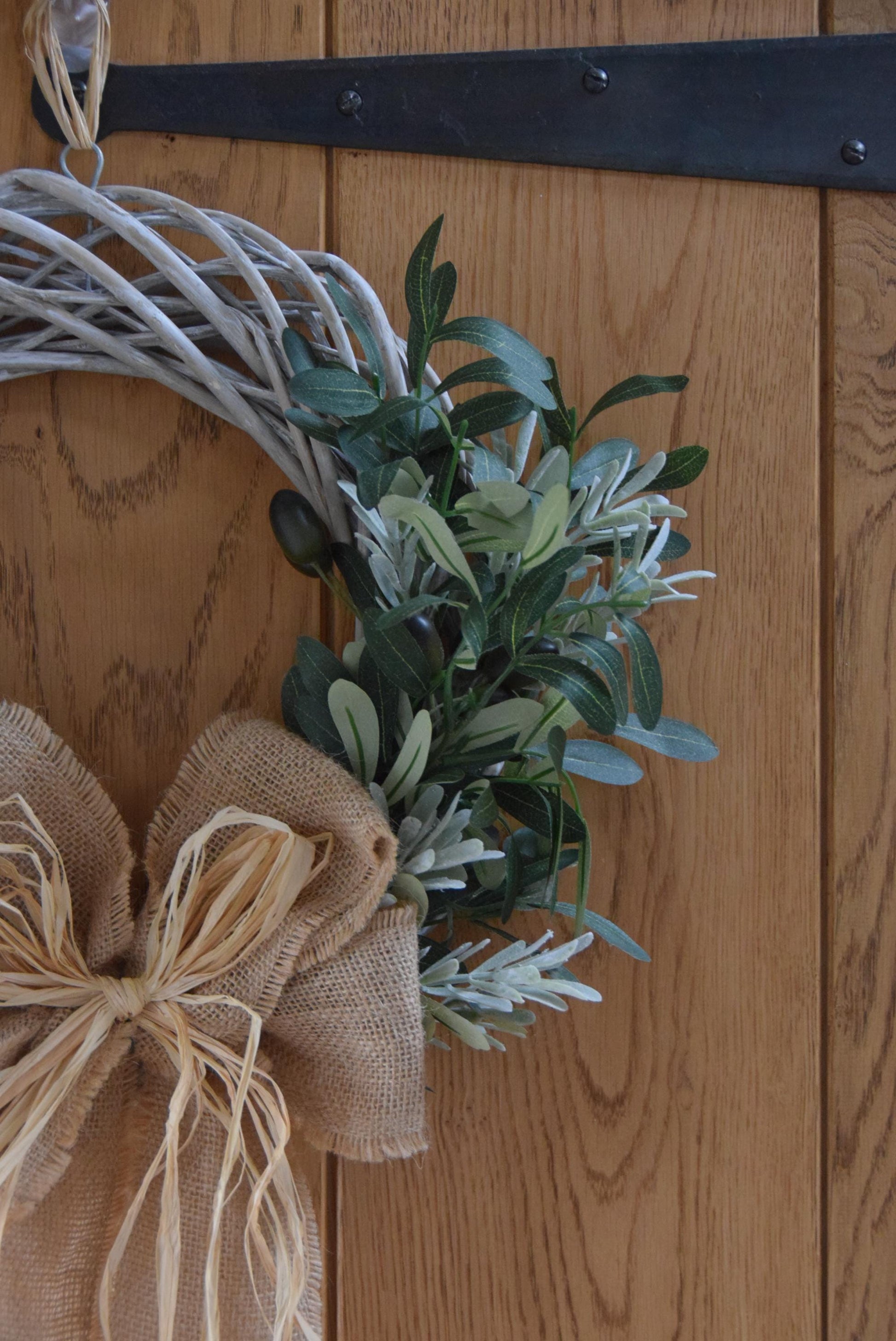 Olive branch wreath created on a willow wreath hanging on a country style door. Mixed with olive branches and tarragon herbs, finished with raffia and natural ribbon.