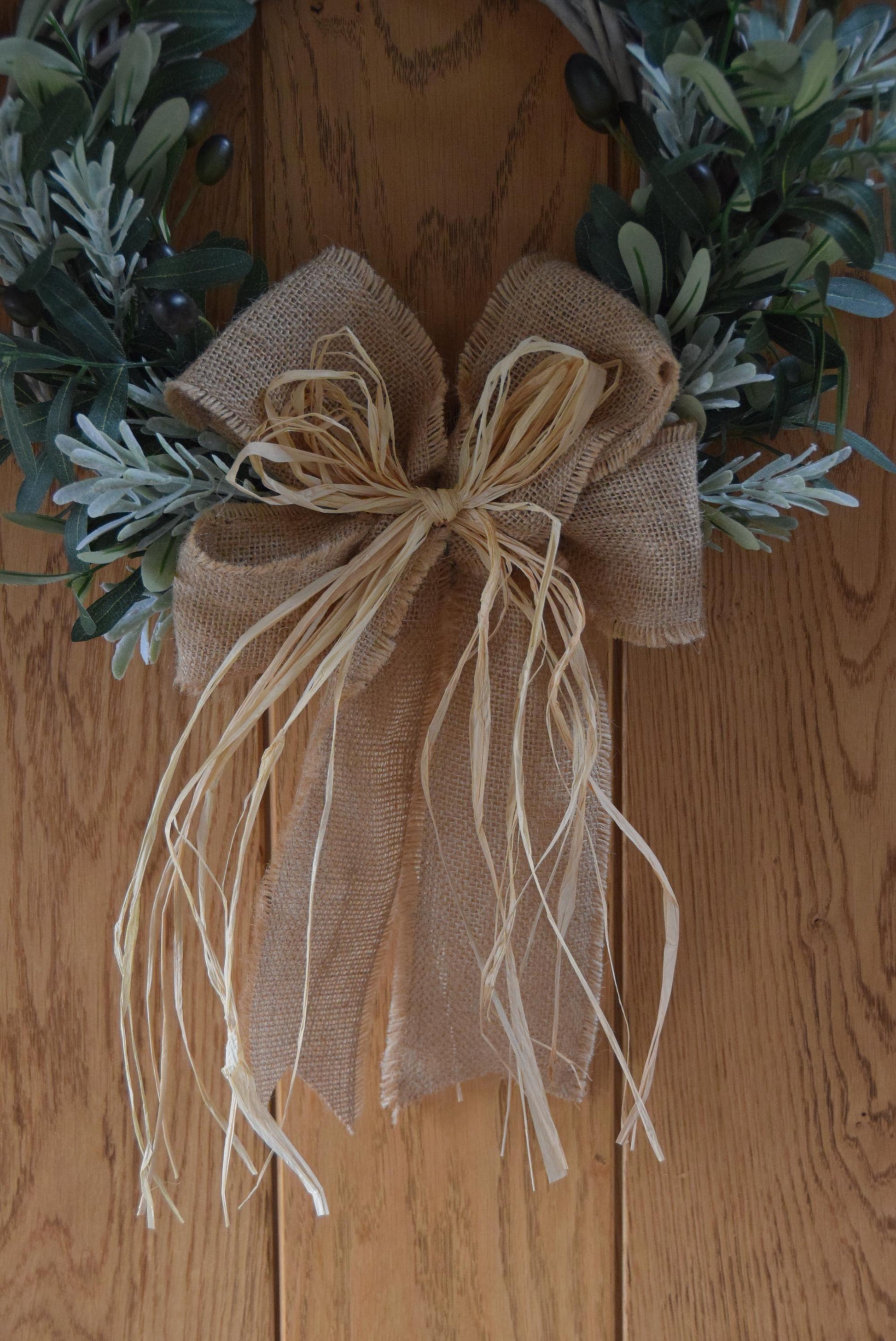 Olive branch wreath created on a willow wreath hanging on a country style door. Mixed with olive branches and tarragon herbs, finished with raffia and natural ribbon.
