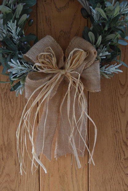 Olive branch wreath created on a willow wreath hanging on a country style door. Mixed with olive branches and tarragon herbs, finished with raffia and natural ribbon.