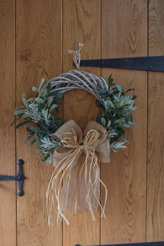Olive branch wreath created on a willow wreath hanging on a country style door. Mixed with olive branches and tarragon herbs, finished with raffia and natural ribbon.