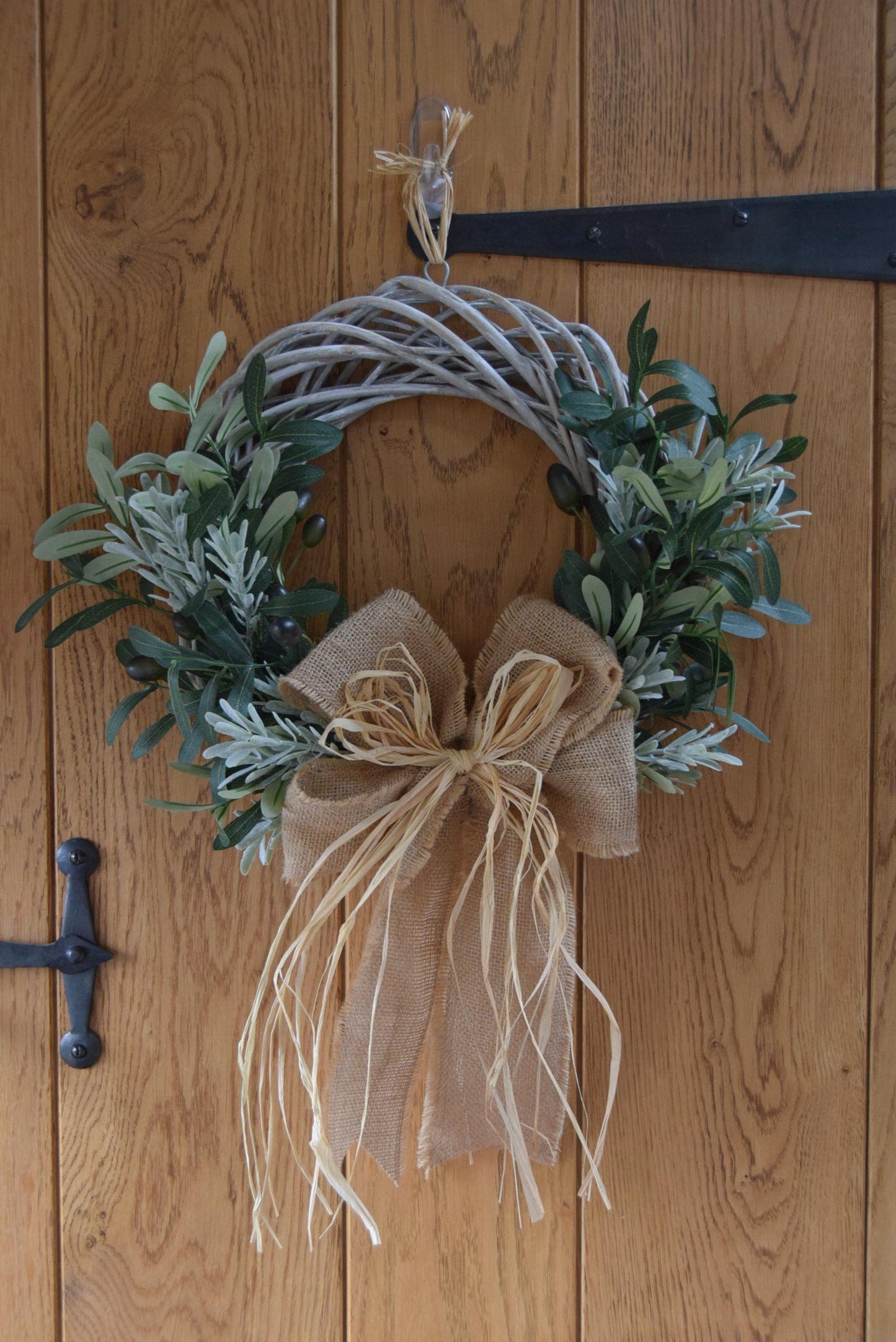 Olive branch wreath created on a willow wreath hanging on a country style door. Mixed with olive branches and tarragon herbs, finished with raffia and natural ribbon.