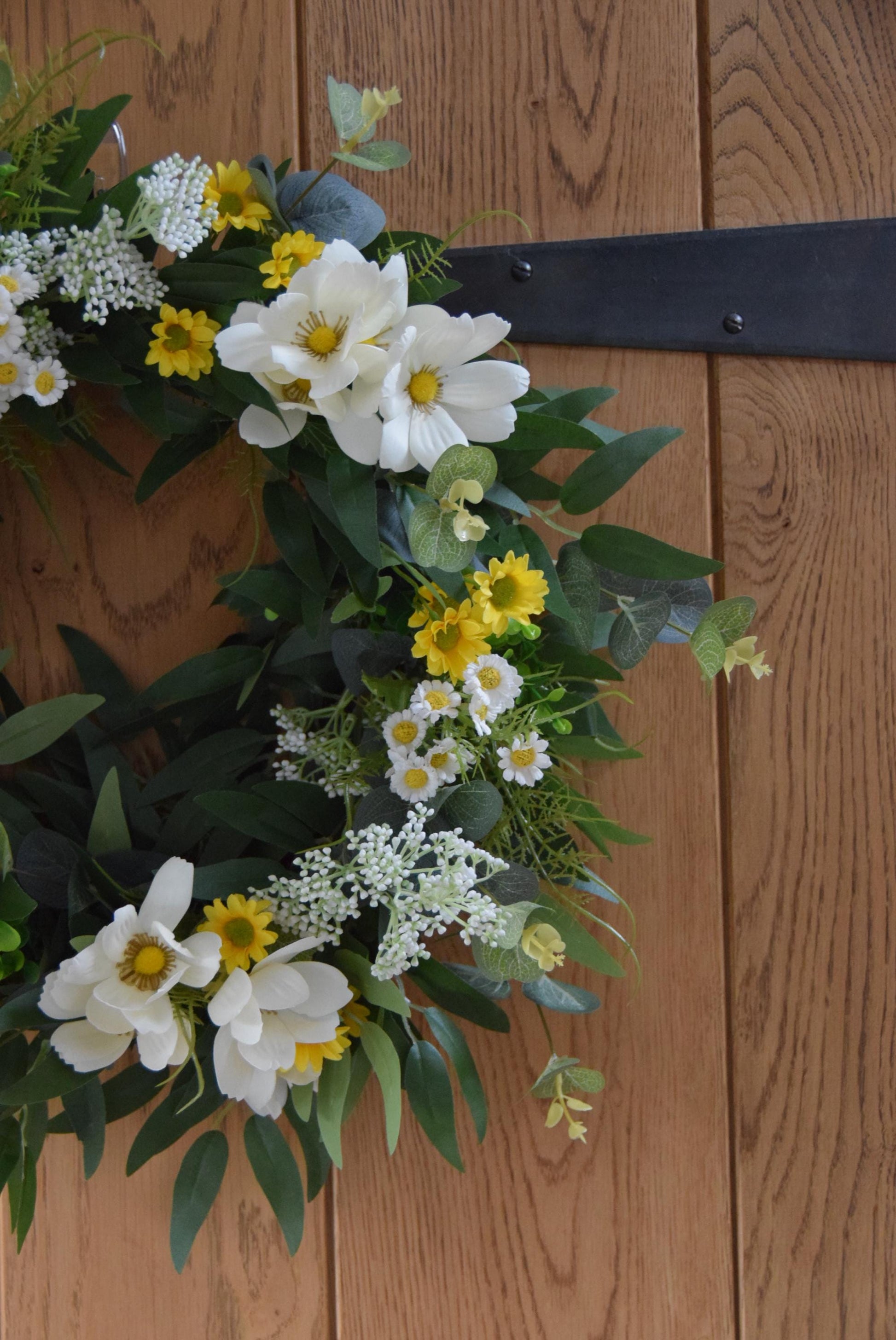 Wreath hanging on rustic country style wooden door with a mixture of daisies, wildflowers, mini bud sprays and greenery. Displayed on a barn door for home decor.