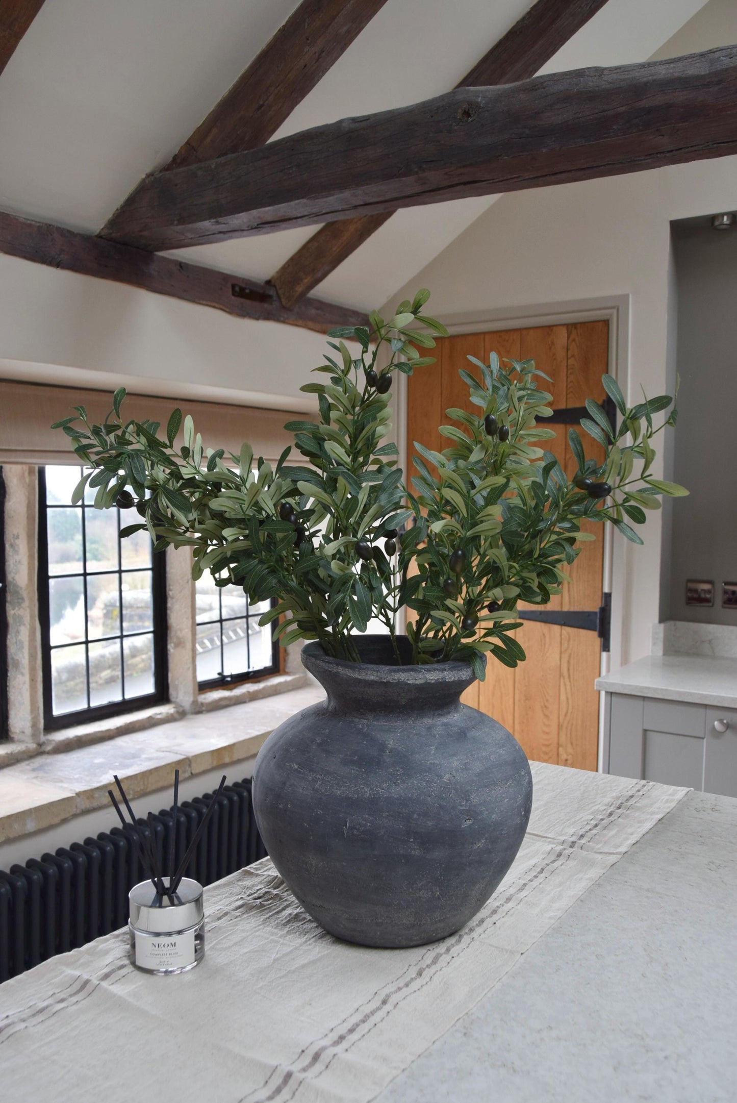 Grey antique vase placed on a kitchen countertop with a bunch of artificial olive branch stems with lots of foliage. Displayed in a country style kitchen for home decor.