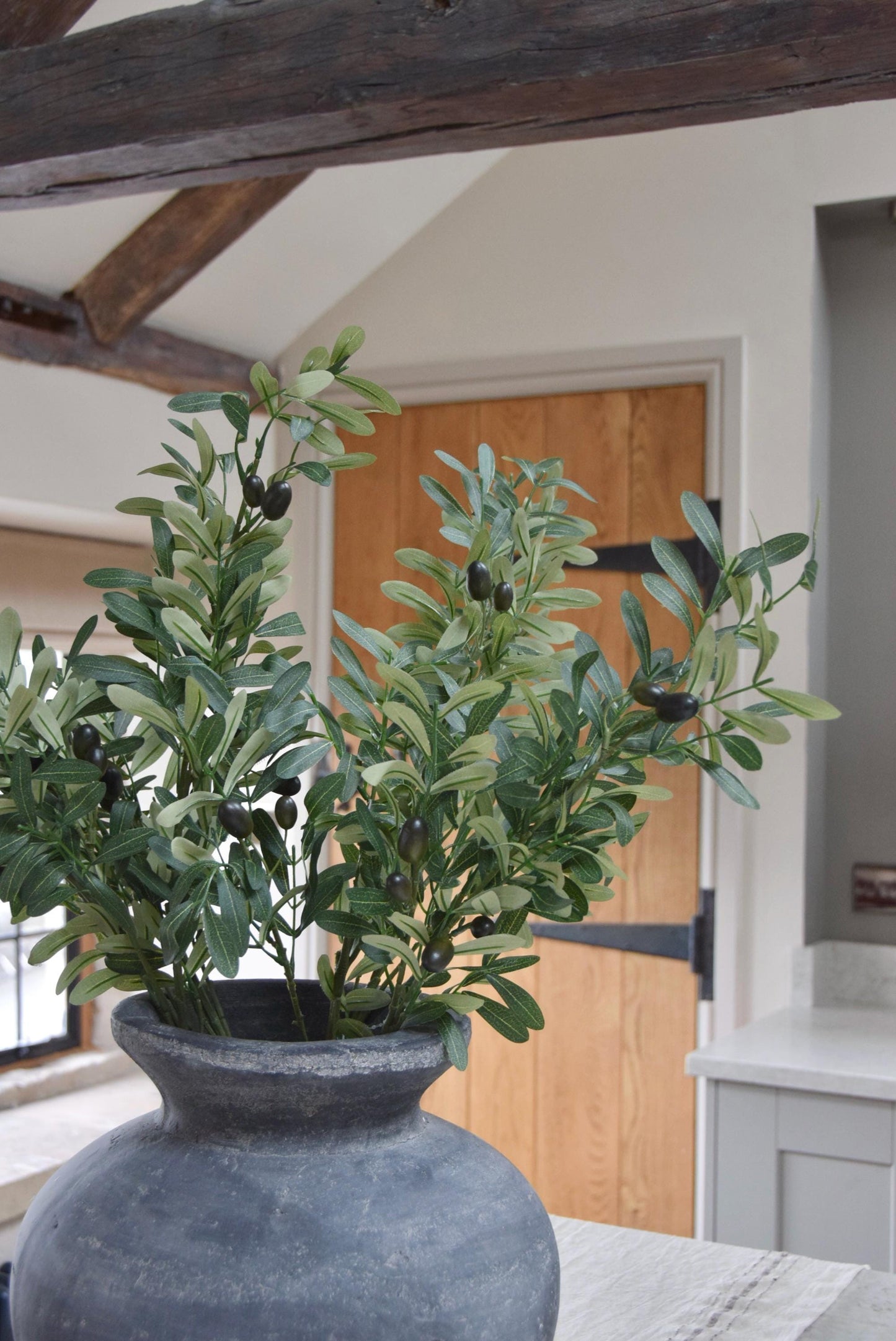 Grey antique vase placed on a kitchen countertop with a bunch of artificial olive branch stems with lots of foliage. Displayed in a country style kitchen for home decor.