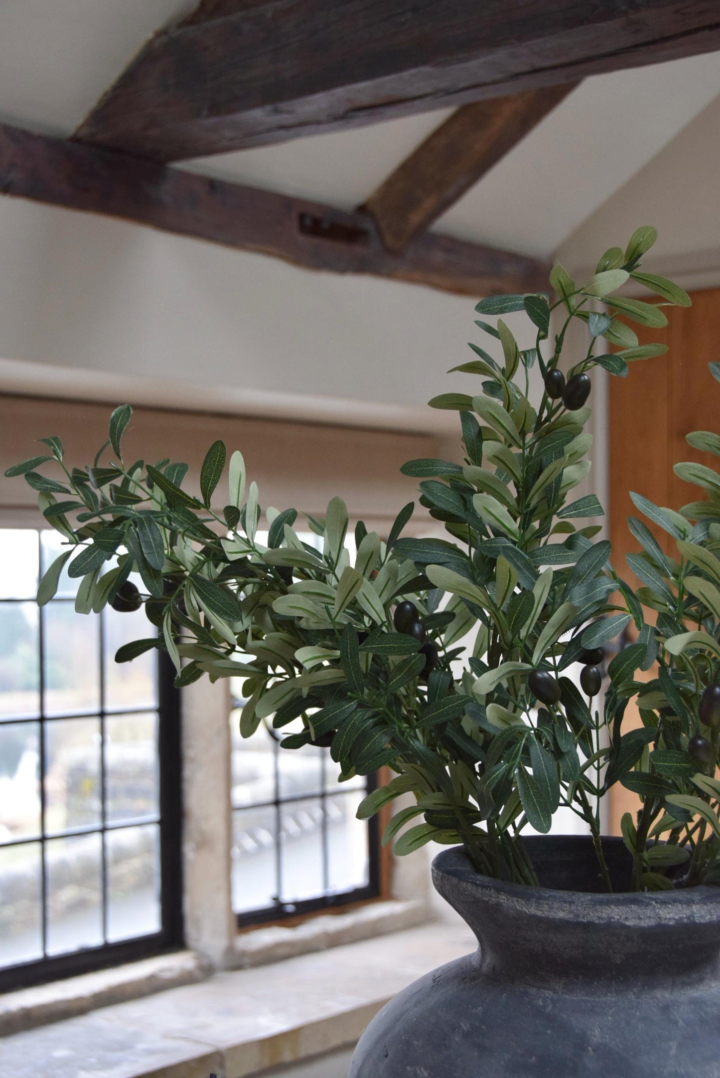 Grey antique vase placed on a kitchen countertop with a bunch of artificial olive branch stems with lots of foliage. Displayed in a country style kitchen for home decor.