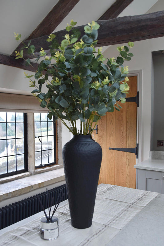 Tall black vase placed on a kitchen countertop with a bunch of green artificial eucalyptus. Displayed in a country style kitchen for home decor.