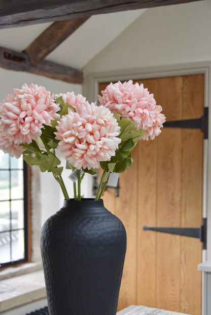 Tall black vase placed on a kitchen counter top with a bunch of artificial pink pom pom flower stems. Displayed in a country style kitchen for home decor.