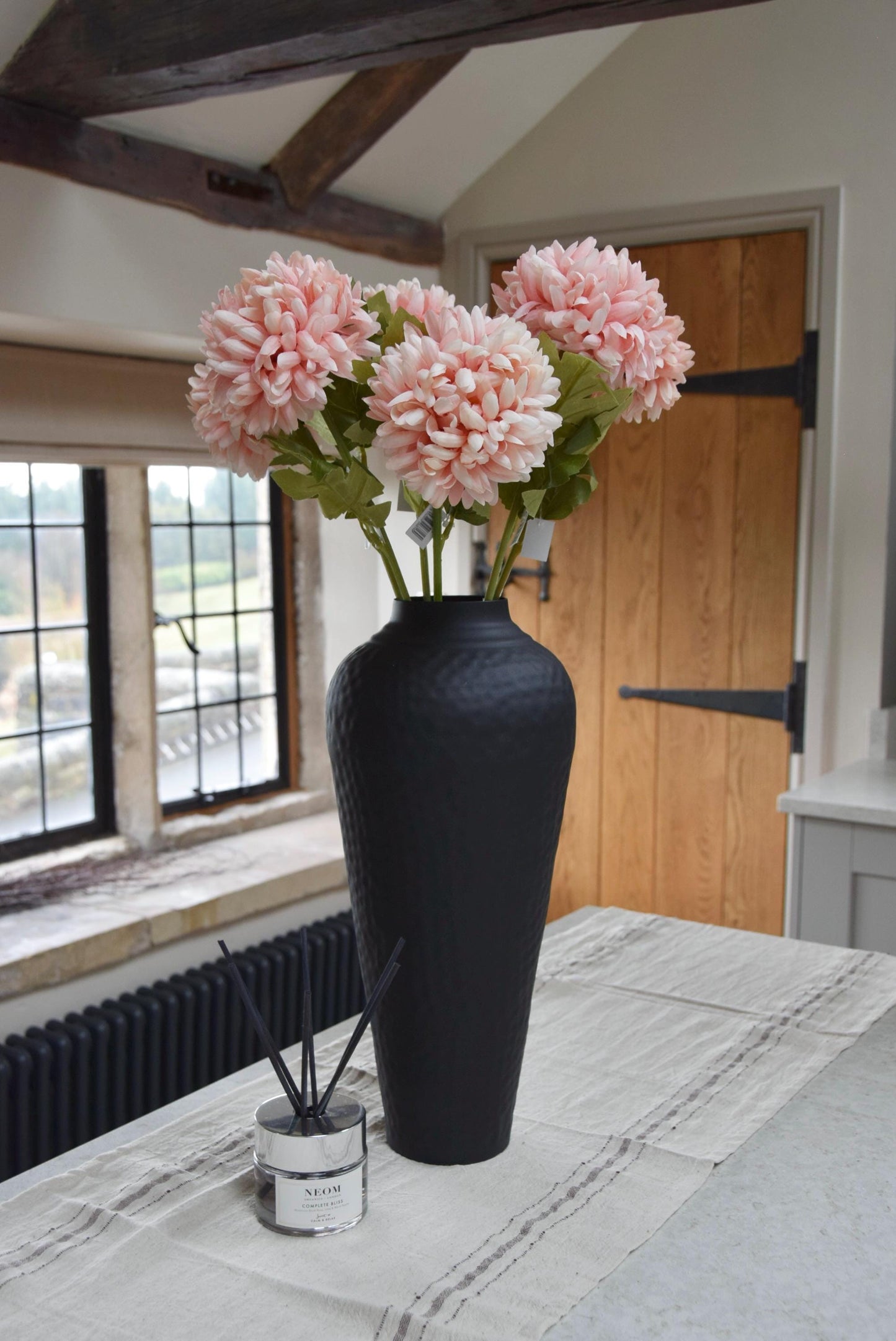 Tall black vase placed on a kitchen counter top with a bunch of artificial pink pom pom flower stems. Displayed in a country style kitchen for home decor.