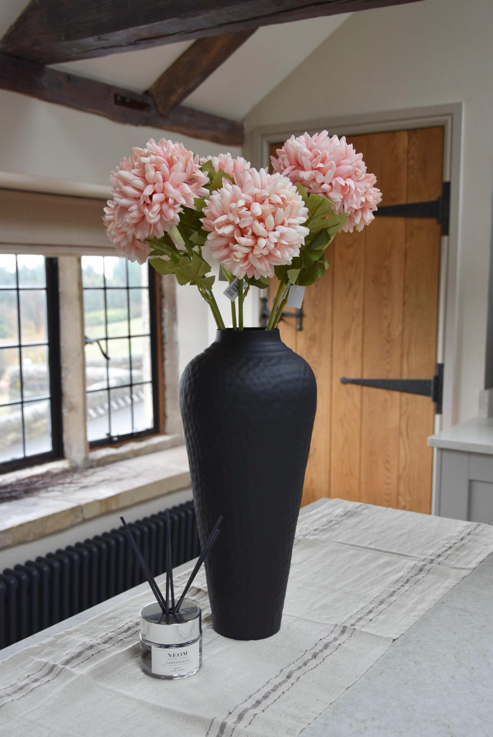 Tall black vase placed on a kitchen counter top with a bunch of artificial pink pom pom flower stems. Displayed in a country style kitchen for home decor.