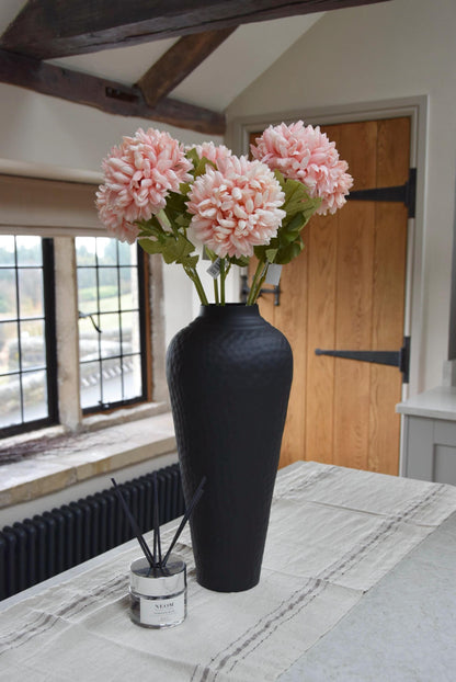 Tall black vase placed on a kitchen counter top with a bunch of artificial pink pom pom flower stems. Displayed in a country style kitchen for home decor.