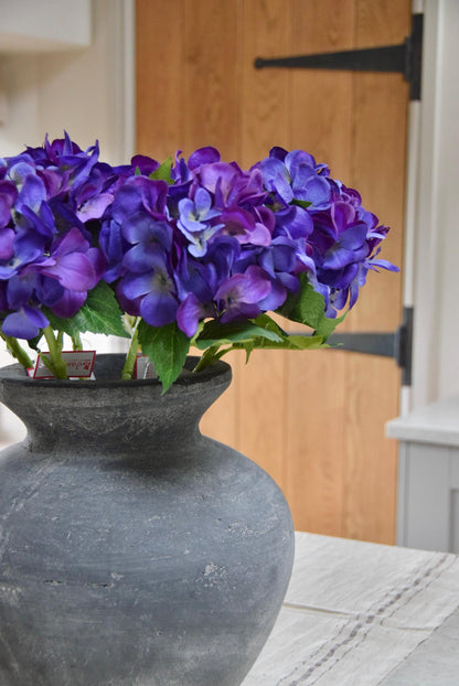 Grey antique vase placed on a kitchen countertop with a bunch of artificial purple hydrangeas. Displayed in a country style kitchen for home decor.
