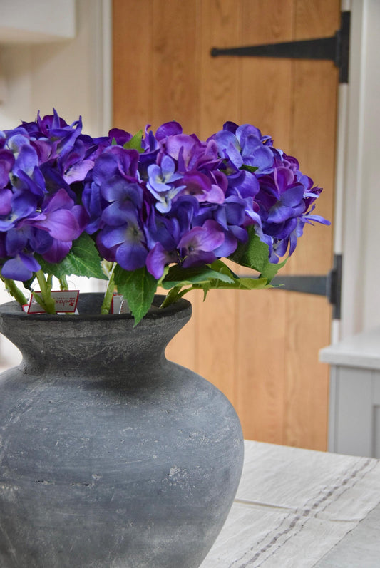 Grey antique vase placed on a kitchen countertop with a bunch of artificial purple hydrangeas. Displayed in a country style kitchen for home decor.