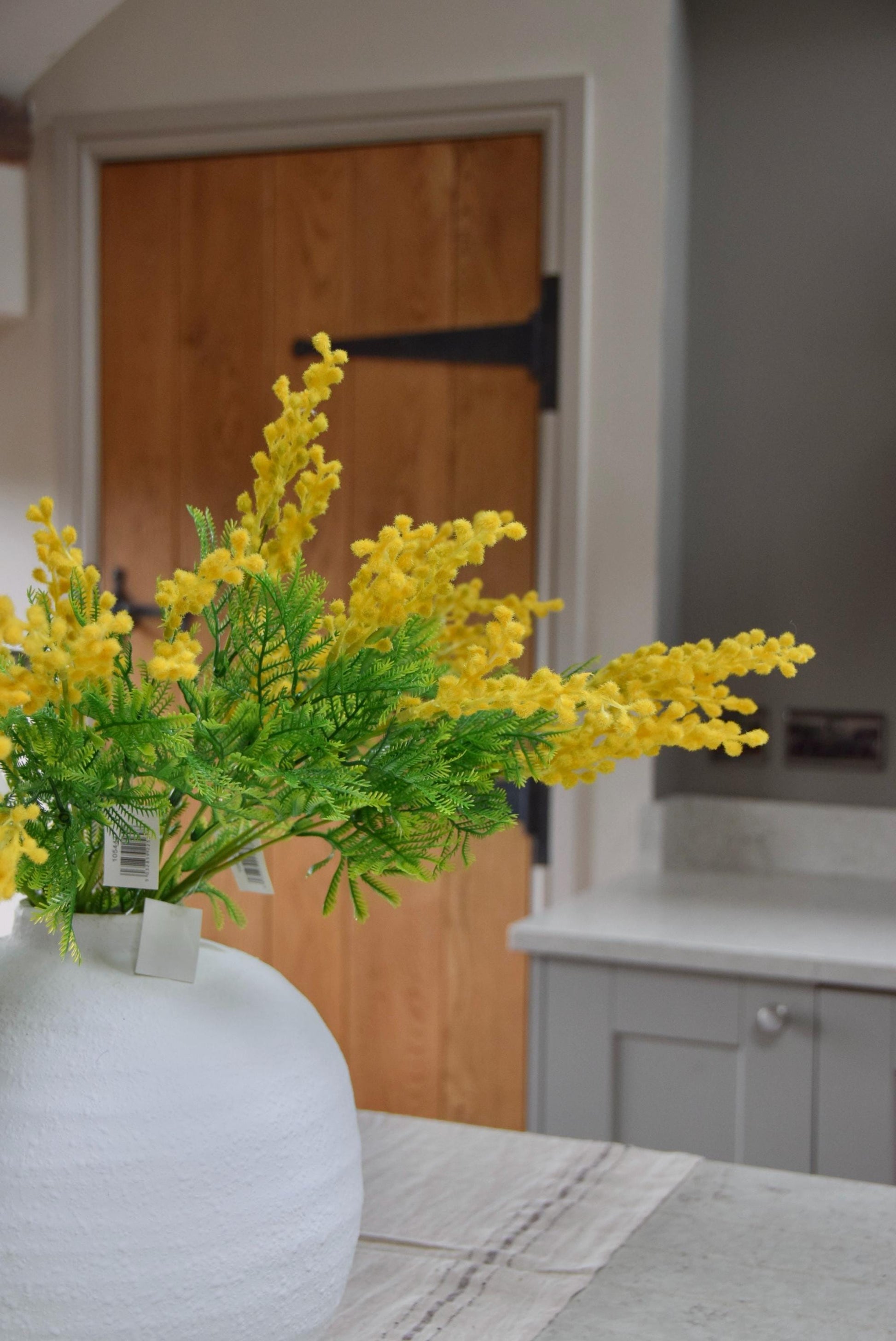 Round white vase placed on a kitchen countertop with a bunch of yellow mimosa stems. Displayed in a country style kitchen for home decor.