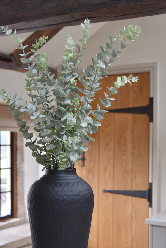 Tall black vase placed on a kitchen countertop with a bunch of dusted artificial eucalyptus stems. Displayed in a country style kitchen for home decor.