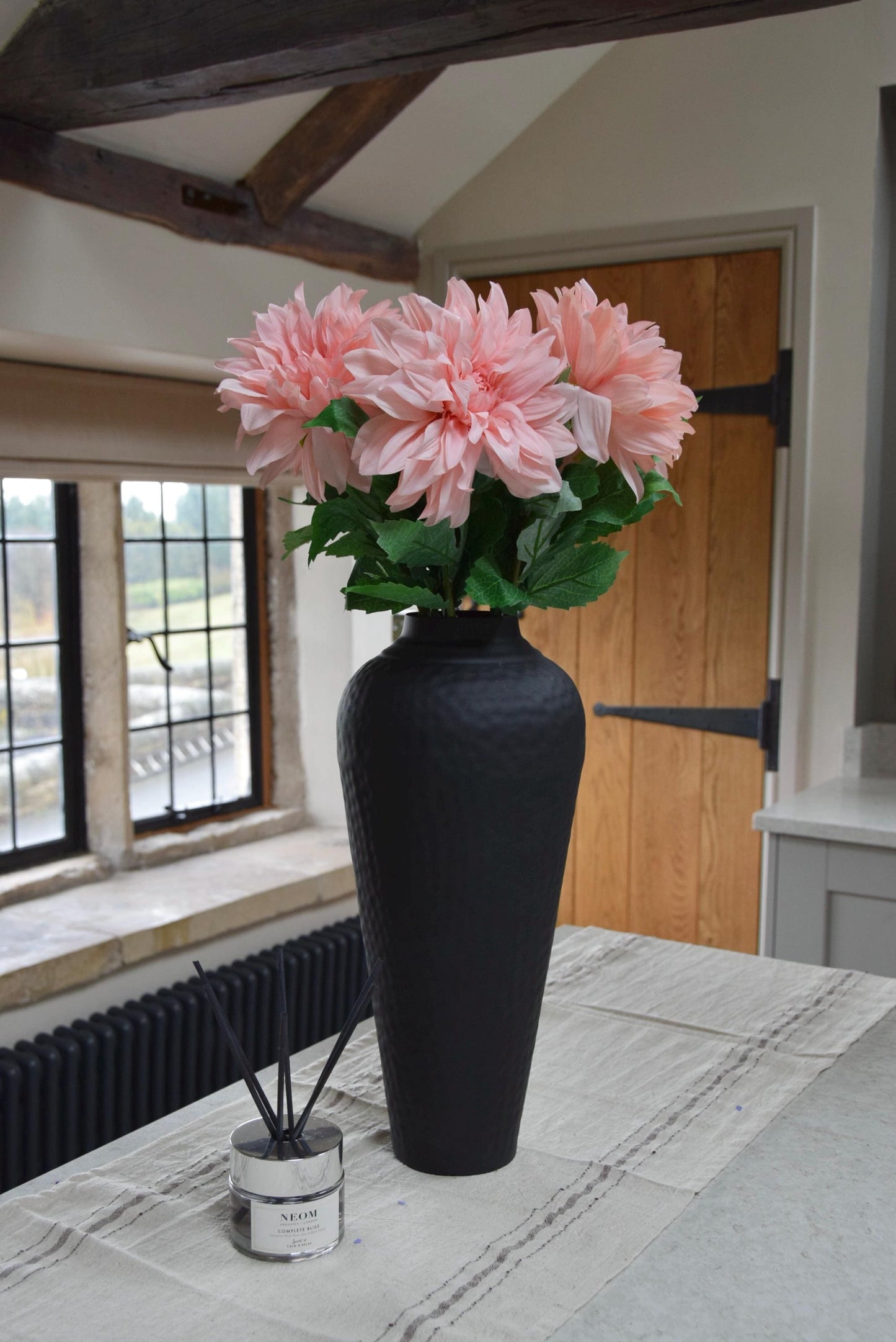 Tall black vase placed on a kitchen countertop with a bunch of pink monarch dahlia stems. Displayed in a country style kitchen for home decor.