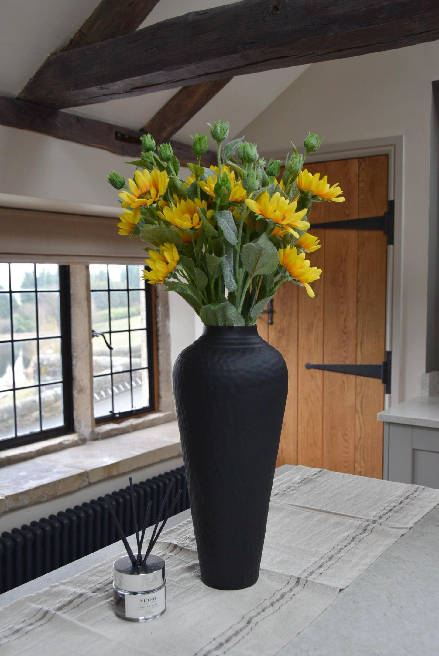 Tall black vase placed on a kitchen countertop with a bunch of artificial yellow sunflower sprays. Displayed in a country style kitchen for home decor.