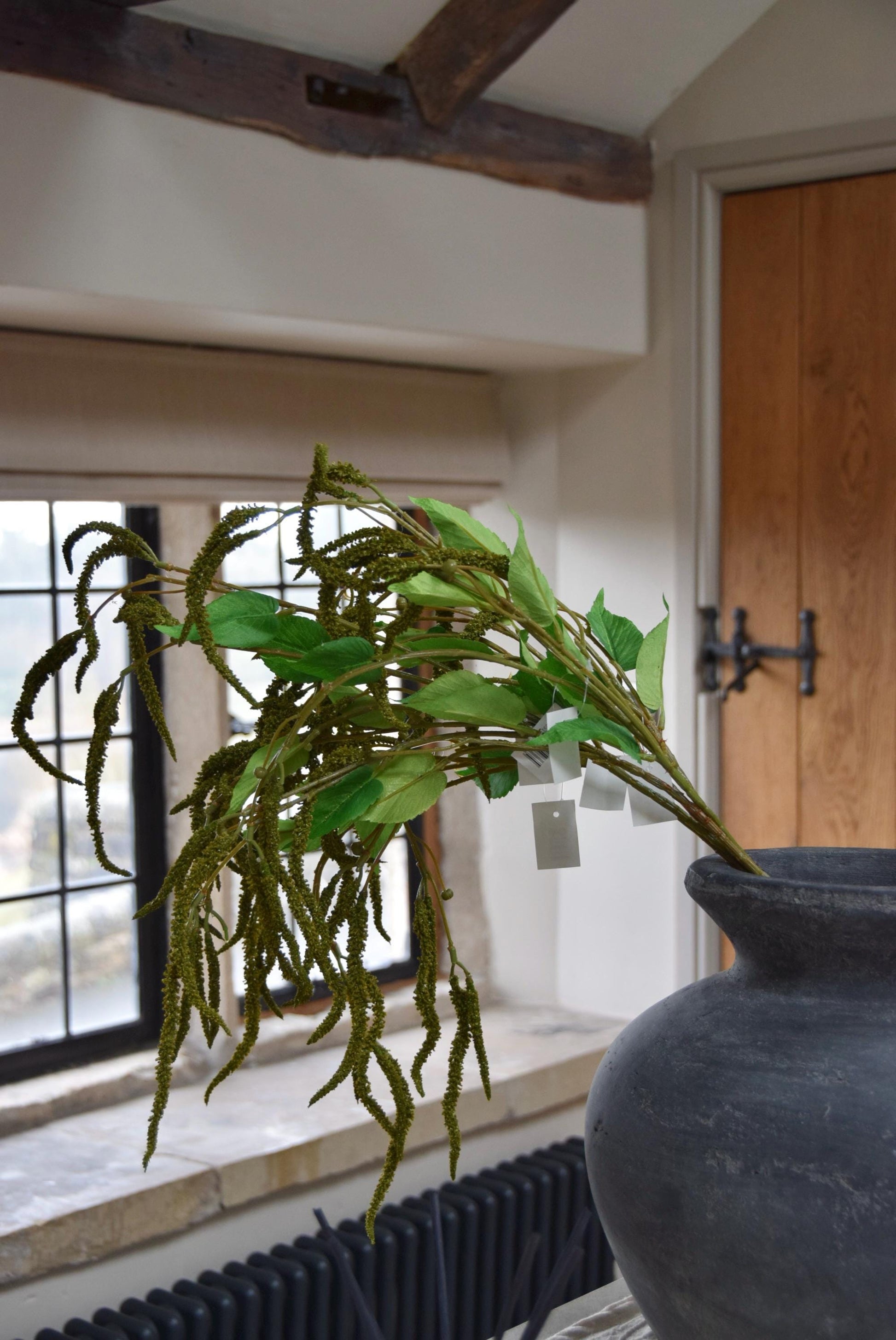 Grey vase placed on a kitchen countertop with bunch of artificial green catkin stems. Displayed in a country style kitchen for home decor.