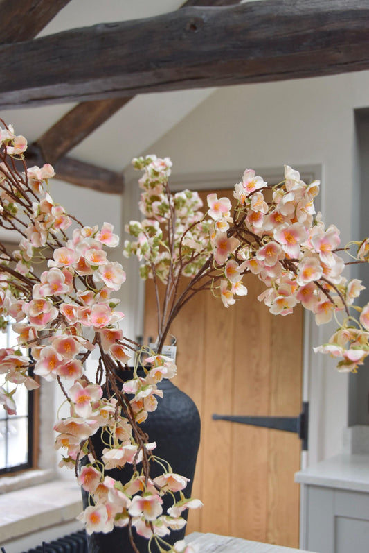 Tall black vase placed on a kitchen countertop with a bunch of hanging pink plum blossom stems. Displayed in a country style kitchen for home decor.