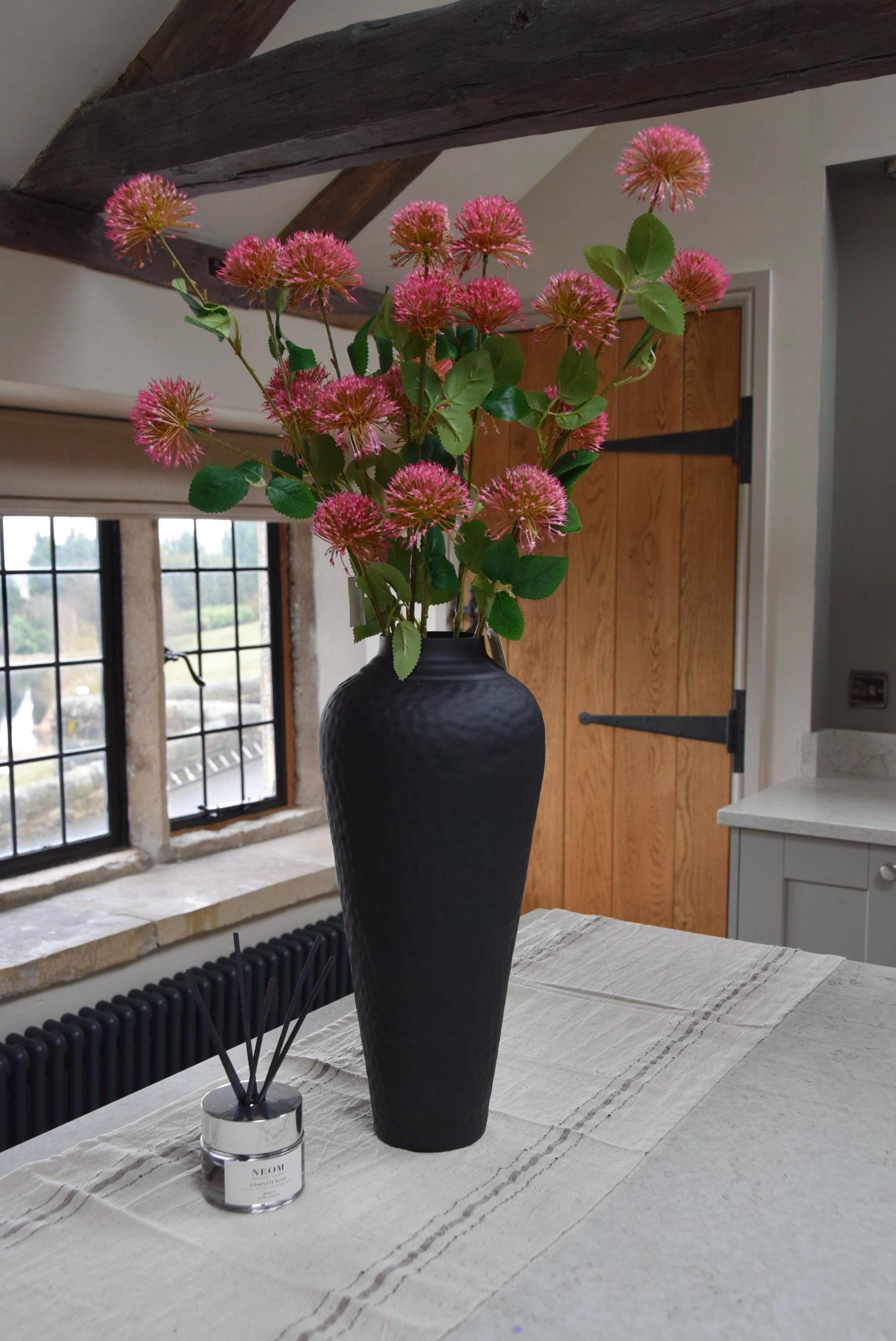Tall black vase placed on a kitchen countertop with a bunch of pink pom flower stems. Displayed in a country style kitchen for home decor.
