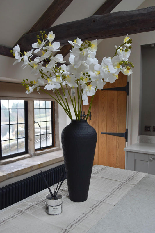 Tall black vase placed on a kitchen counter top with a bunch of white orchid sprays. Displayed in a country style kitchen for home decor.