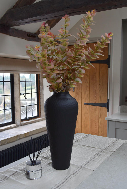 Tall black vase placed on a kitchen countertop with a bunch of pink and green blush eucalyptus stems. Displayed in a country style kitchen for home decor.