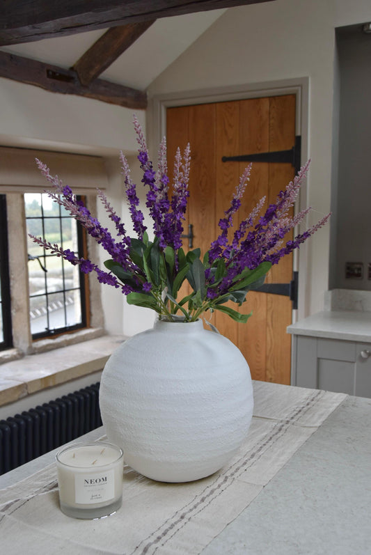 White round vase with a bunch of purple wild flower stems placed on a kitchen countertop. Displayed in a country style kitchen for home decor.