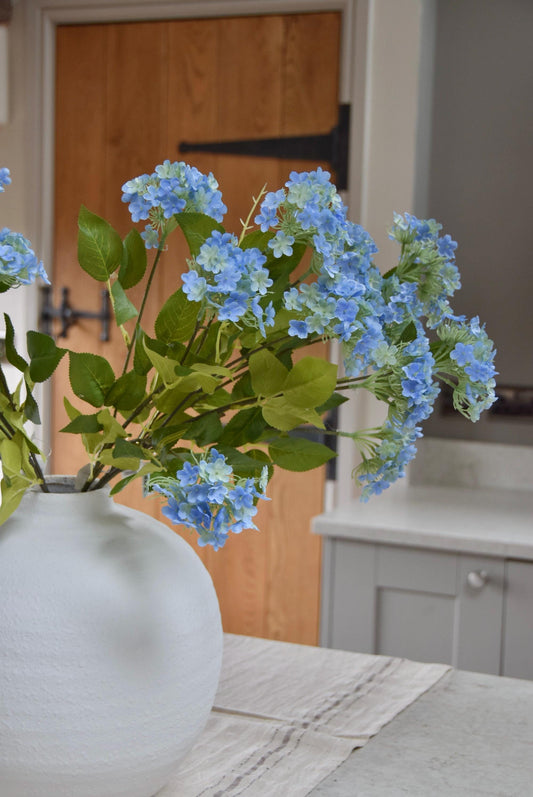 White round vase placed on a kitchen countertop with a bunch of blue wildflower stems displayed in a country style kitchen for home decor.