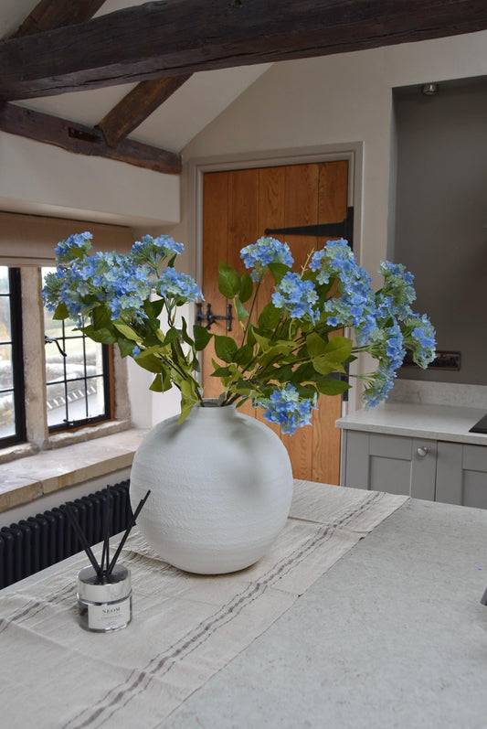 White round vase placed on a kitchen countertop with a bunch of blue wildflower stems displayed in a country style kitchen for home decor.
