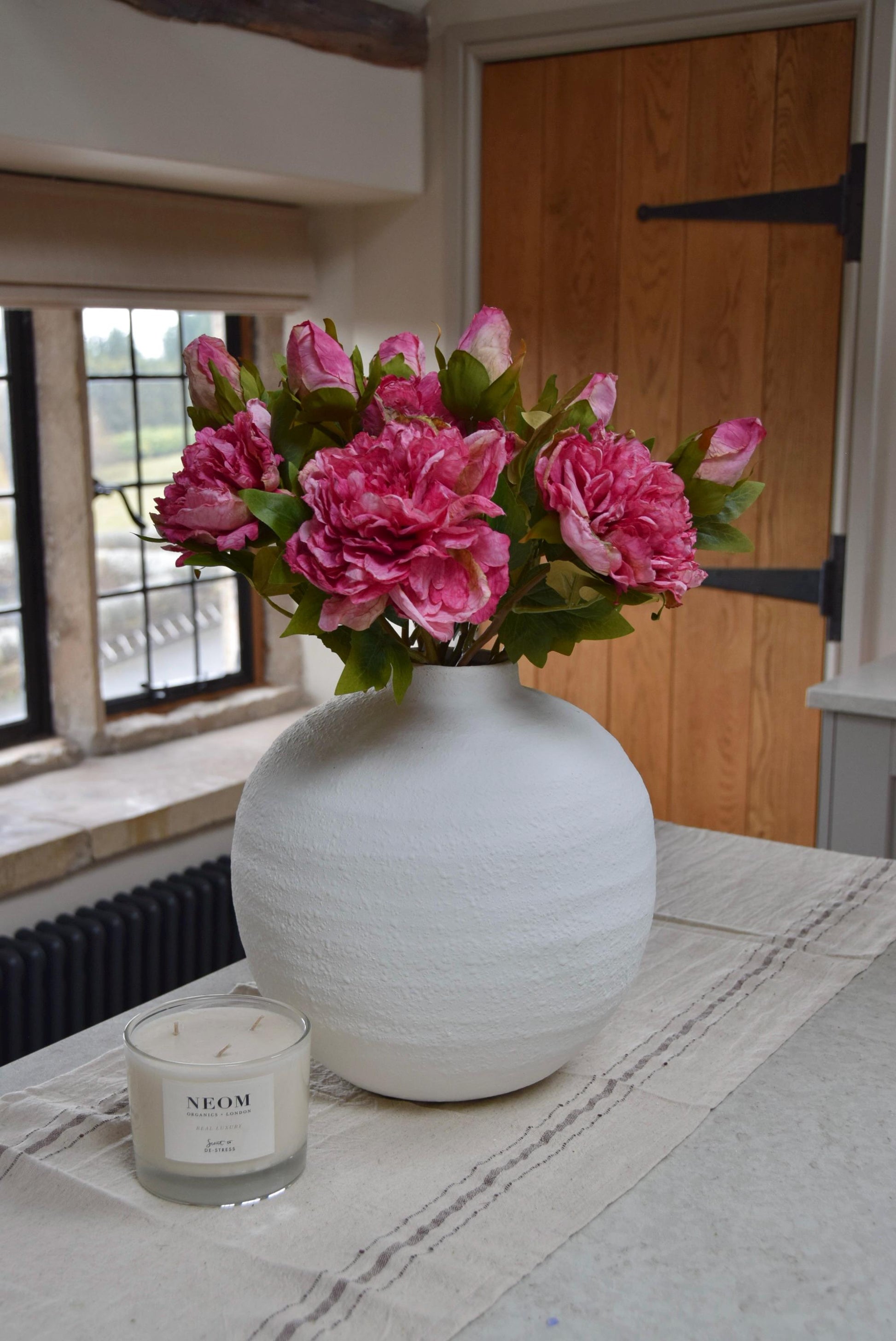 White round vase placed on a kitchen countertop with a bunch of hot pink peonies. Displayed in a country style kitchen for home decor.