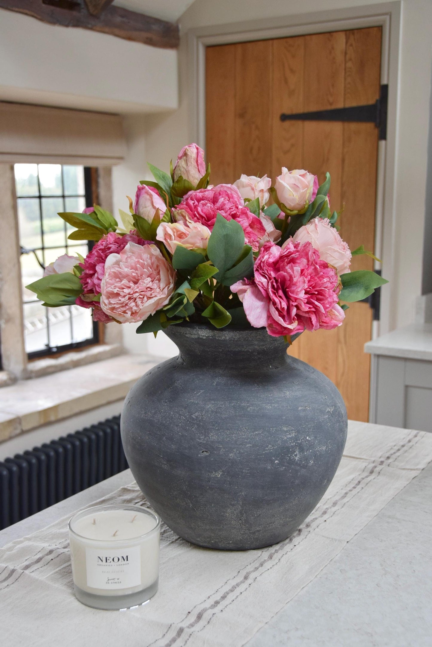 Grey antique vase placed on kitchen counter top with an arrangement of pale pink and hot pink peonies. Arranged in a country style kitchen for home decor.
