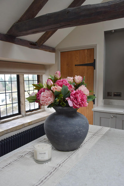 Grey antique vase placed on kitchen counter top with an arrangement of pale pink and hot pink peonies. Arranged in a country style kitchen for home decor.