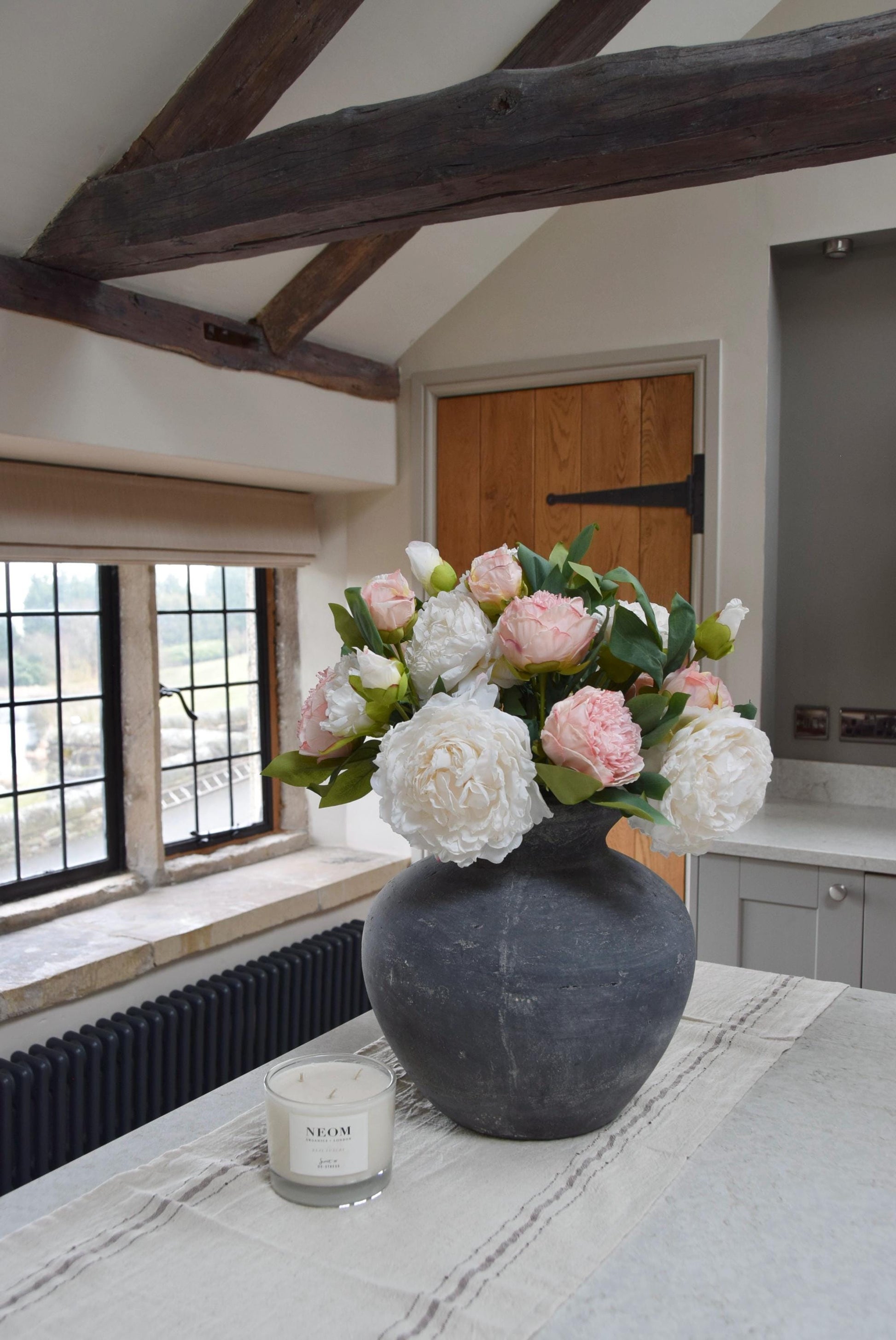 Grey antique vase placed on a kitchen counter top with an arrangement of pink and cream peonies. Arranged in a country style kitchen for home decor.