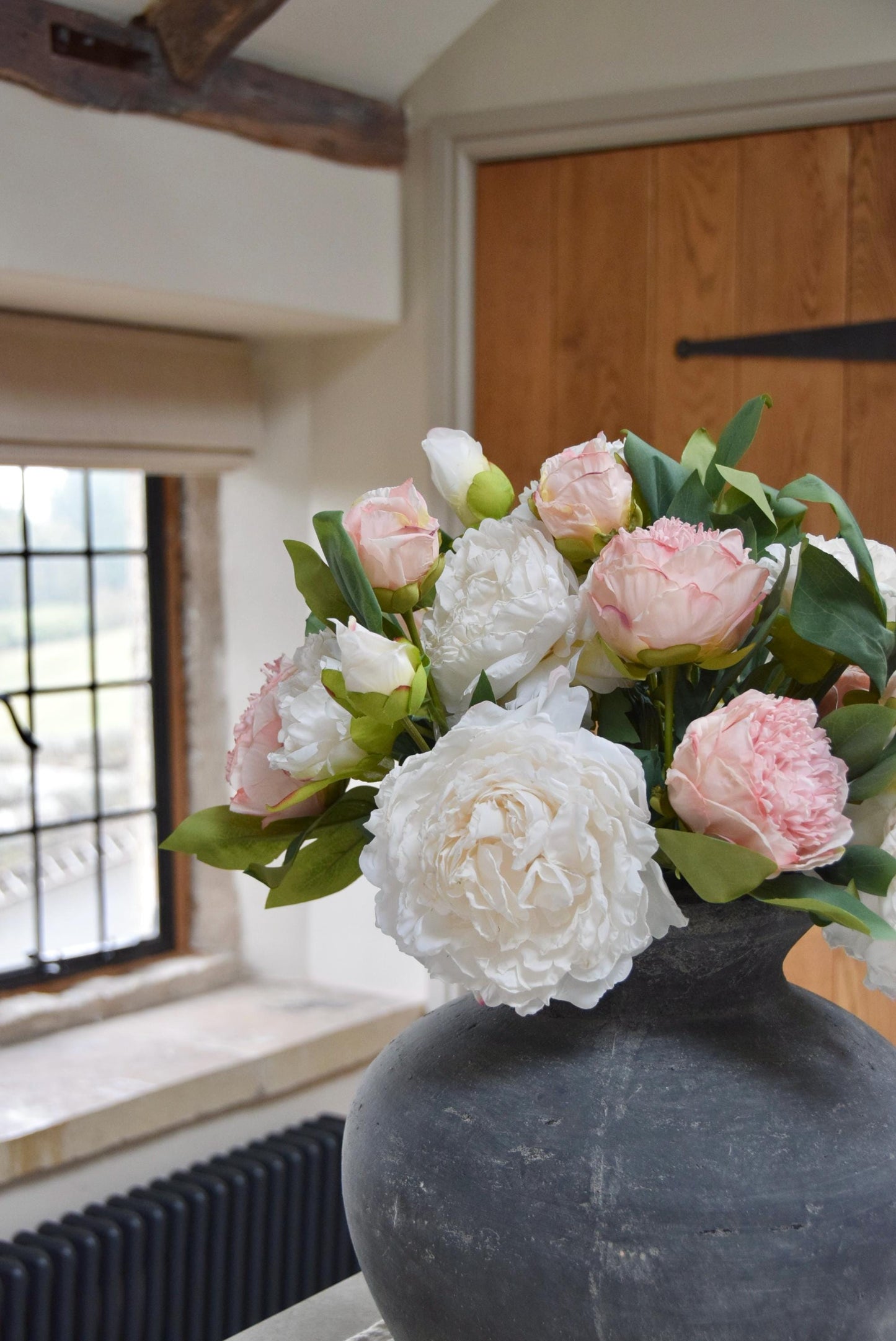 Grey antique vase placed on a kitchen counter top with an arrangement of pink and cream peonies. Arranged in a country style kitchen for home decor.