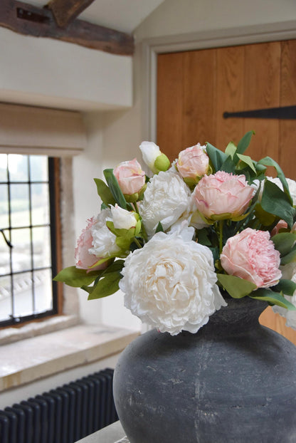 Grey antique vase placed on a kitchen counter top with an arrangement of pink and cream peonies. Arranged in a country style kitchen for home decor.