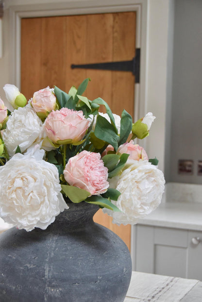 Grey antique vase placed on a kitchen counter top with an arrangement of pink and cream peonies. Arranged in a country style kitchen for home decor.