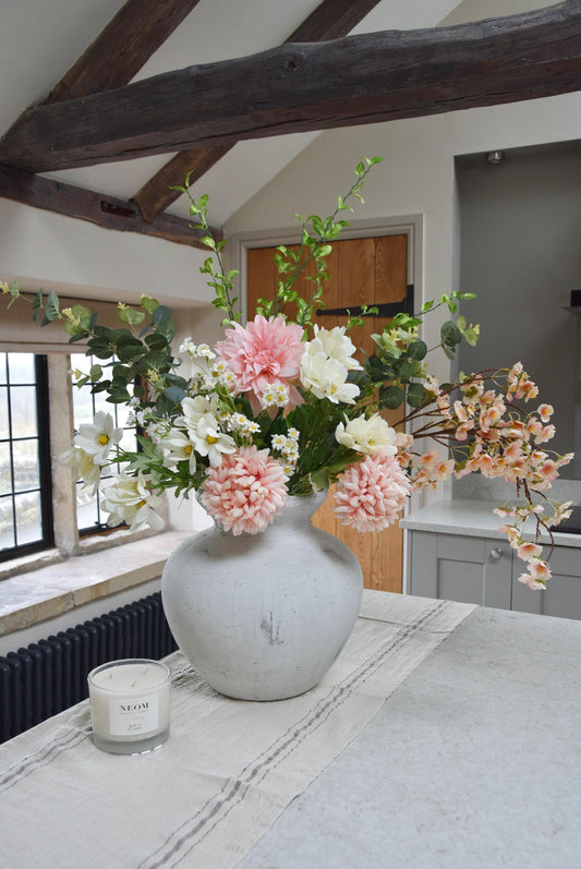 Stone antique vase placed on a kitchen counter top with an arrangement of pink dahlias and mum flowers with white wildflowers and green foliage. Set in a country style kitchen for home decor.