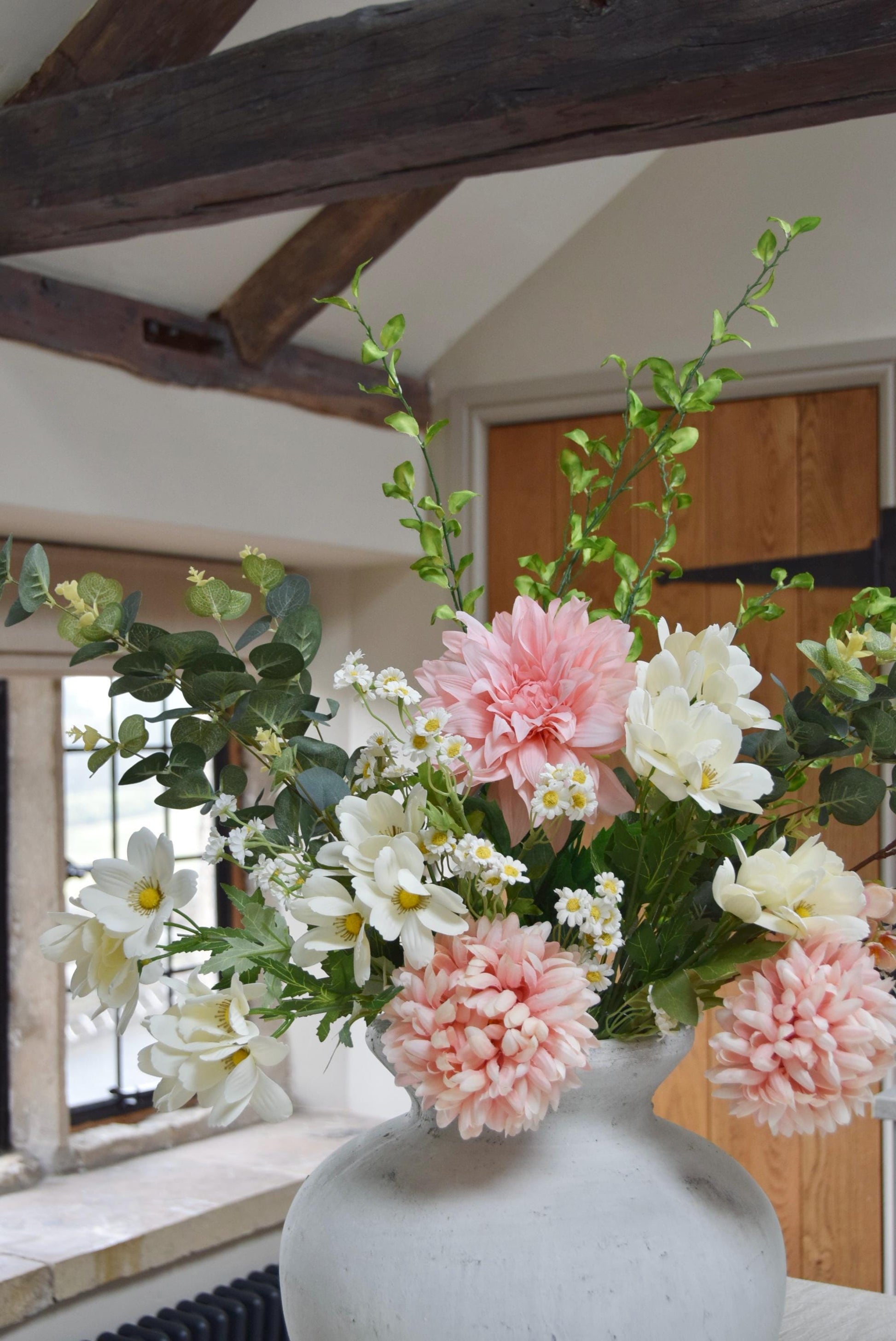 Stone antique vase placed on a kitchen counter top with an arrangement of pink dahlias and mum flowers with white wildflowers and green foliage. Set in a country style kitchen for home decor.