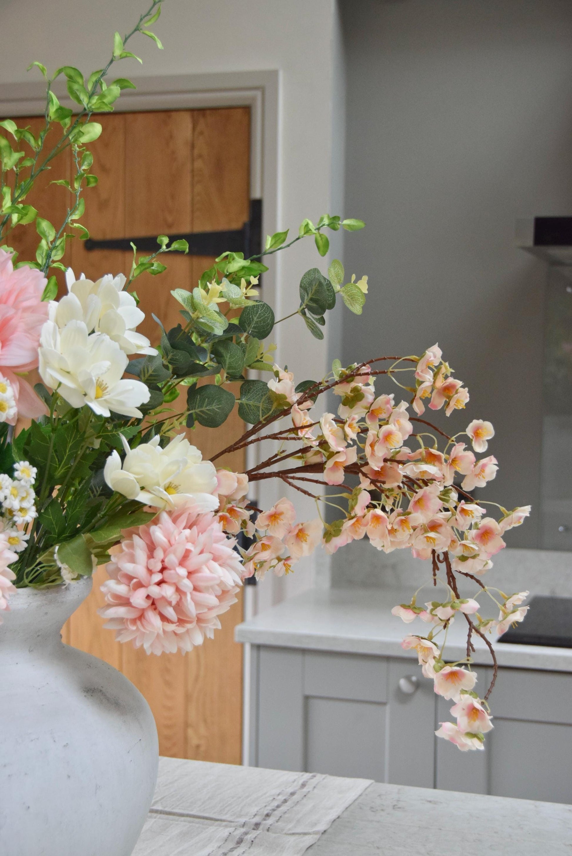 Stone antique vase placed on a kitchen counter top with an arrangement of pink dahlias and mum flowers with white wildflowers and green foliage. Set in a country style kitchen for home decor.