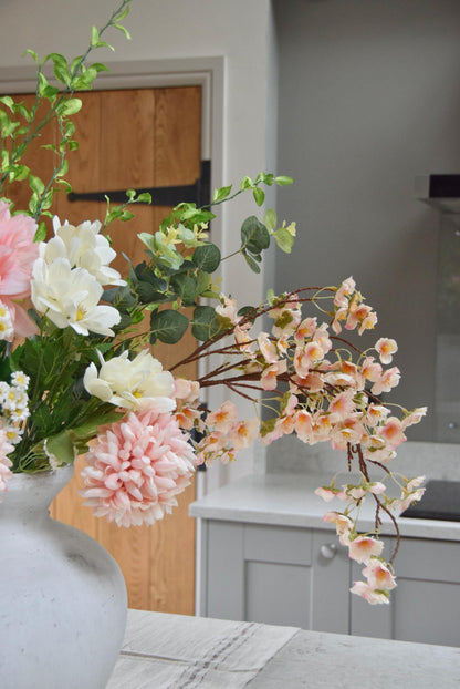 Stone antique vase placed on a kitchen counter top with an arrangement of pink dahlias and mum flowers with white wildflowers and green foliage. Set in a country style kitchen for home decor.
