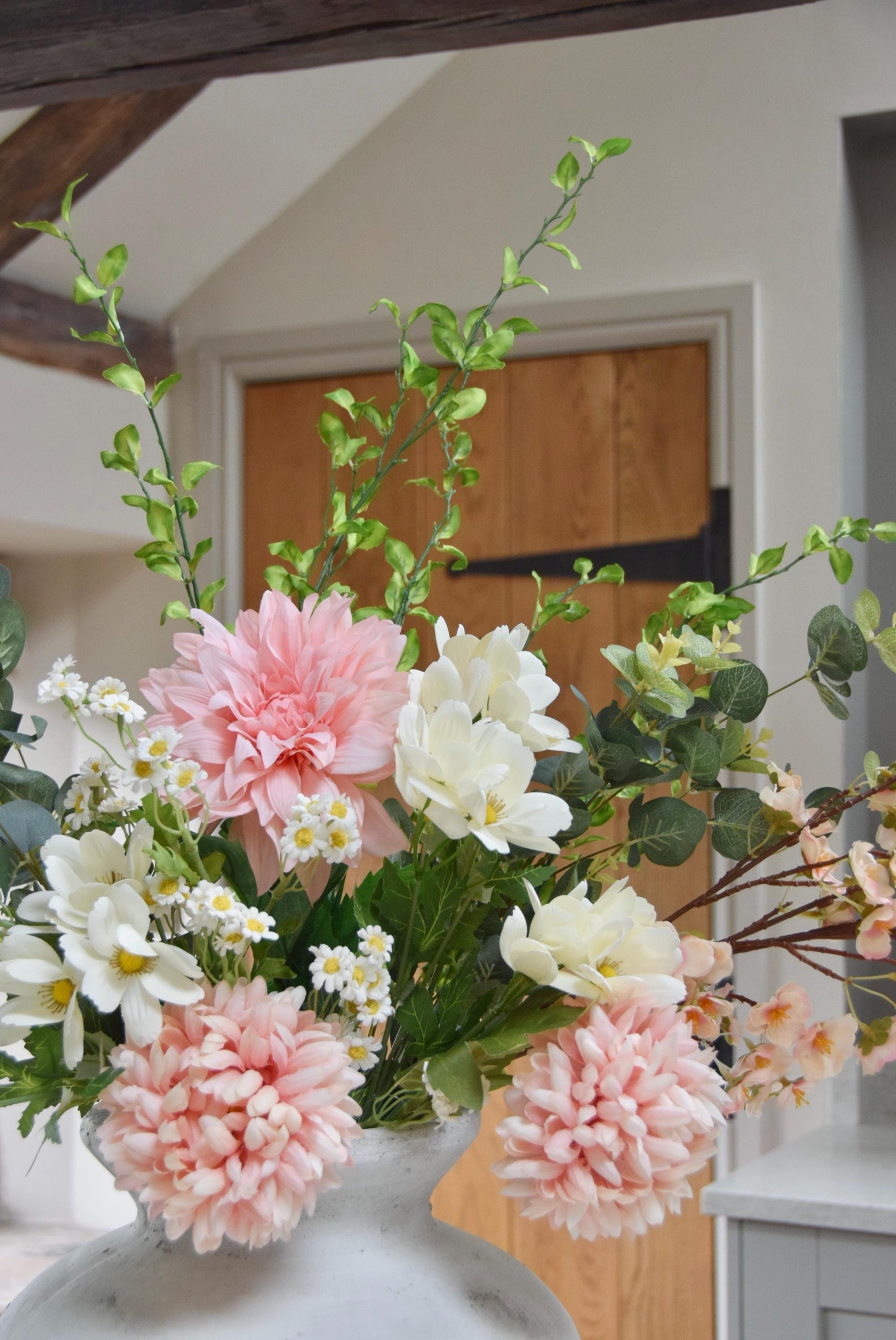 Stone antique vase placed on a kitchen counter top with an arrangement of pink dahlias and mum flowers with white wildflowers and green foliage. Set in a country style kitchen for home decor.