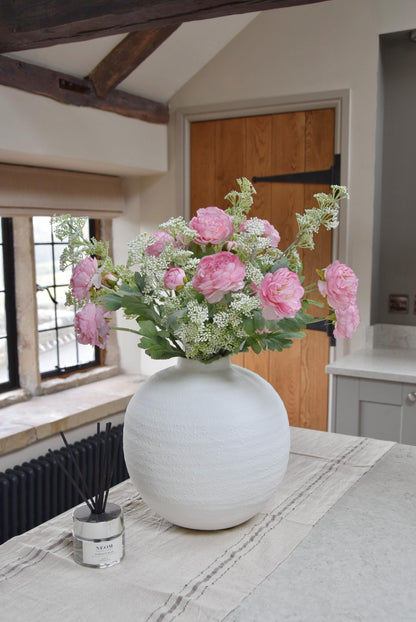 White sphere vase placed on a kitchen countertop with an arrangement of pale pink ranunculuses and white mini bud stems. Displayed in a country style kitchen for home decor.